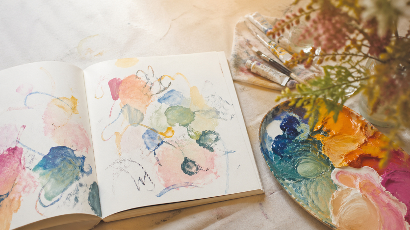 抽象的な色と線が自由に描かれたスケッチブックと、色鮮やかな絵の具のパレット