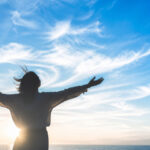 青空の下で両手を広げ、心地よい風を受けながら立つ人物のシルエット