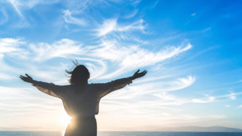 青空の下で両手を広げ、心地よい風を受けながら立つ人物のシルエット